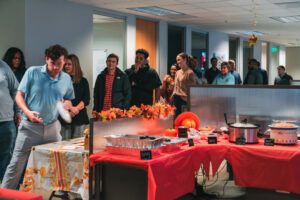 Co-workers chatting and laughing as they serve themselves from a variety of Thanksgiving dishes, including roast vegetables, stuffing, and cranberry sauce.